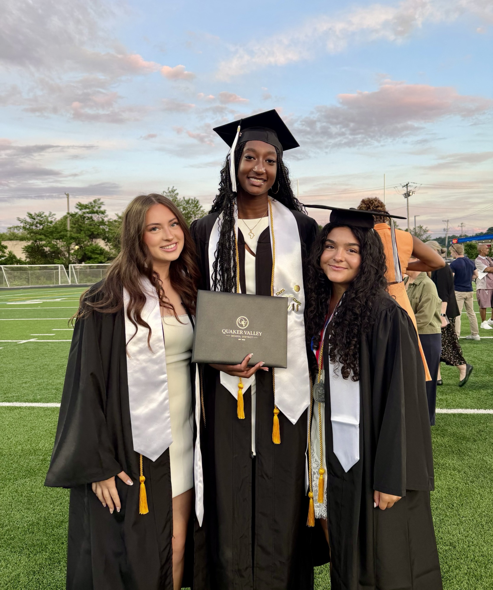 Three QV Girls Basketball seniors in cap and gown at graduation, holding a Quaker Valley diploma at sunset