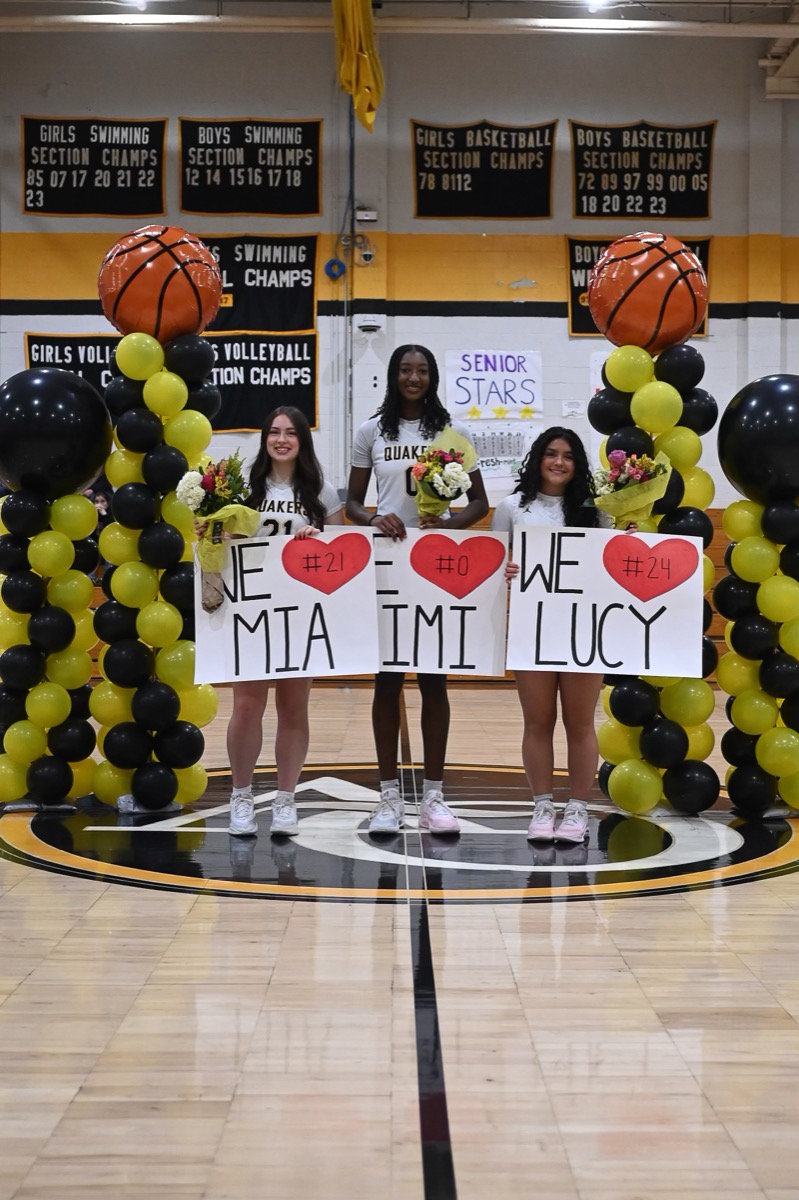Seniors Mia, Mimi, and Lucy holding signs on Senior Night with black and gold balloons